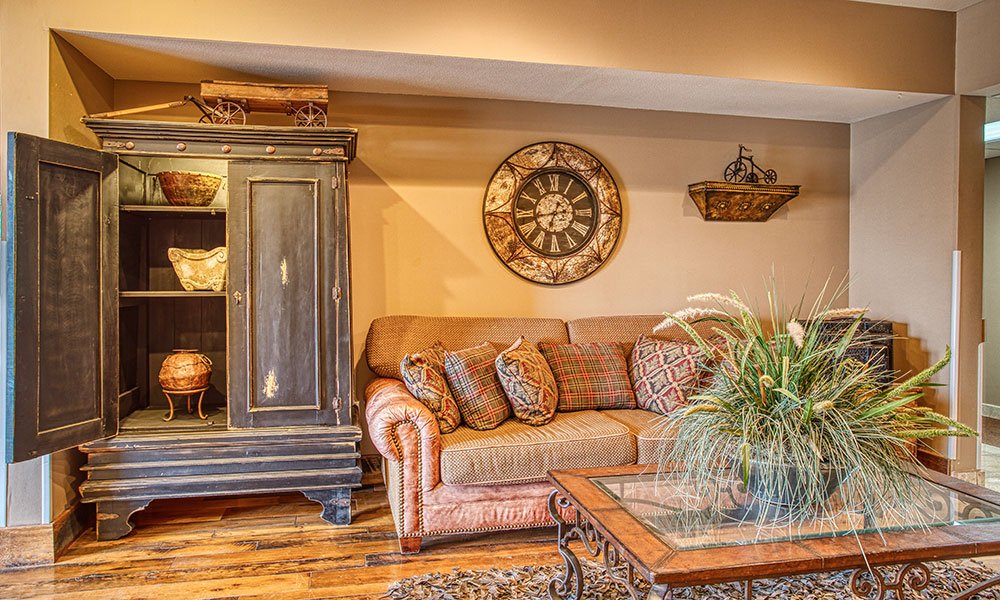 Cozy living room with a tan sofa, plaid pillows, a black armoire with decor inside, a glass coffee table with a plant, a rustic wall clock, and warm brown walls and wood flooring.