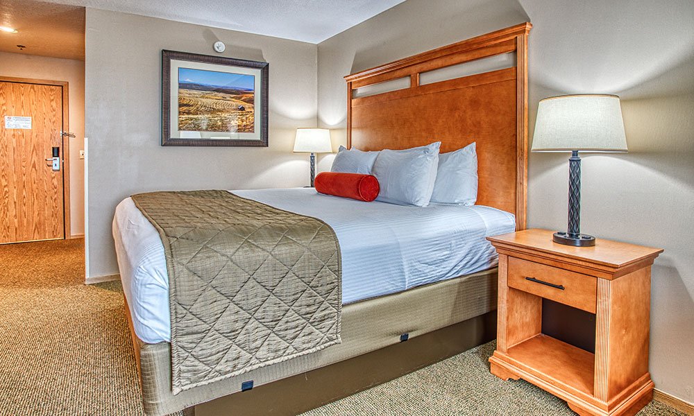 Hotel room with a neatly made king bed, white linens, and a green quilt. Wooden headboard, nightstand, and lamp beside the bed. A framed landscape photo hangs on a beige wall. Door visible in the background.